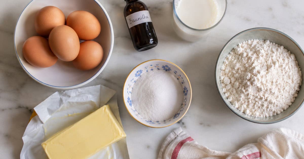 Vista de cima dos ingredientes do bolo amanteigado dispostos em bancada de mármore branco: ovos, farinha, manteiga, baunilha, leite e açúcar