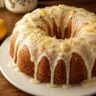 Bolo de limão fofinho com cobertura de cream cheese escorrendo pelas laterais, decorado com raspas de limão, servido em prato branco sobre mesa de madeira