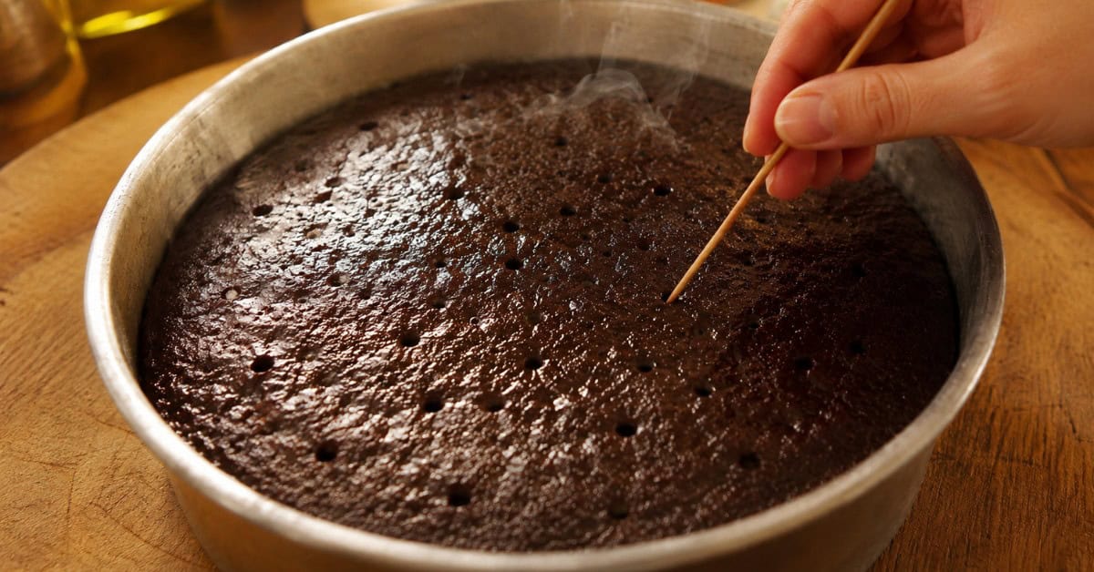 Mãos fazendo furos em bolo de chocolate ainda quente na forma para receber a calda da Nega Maluca sem açúcar, com luz dourada natural e bancada rústica ao fundo