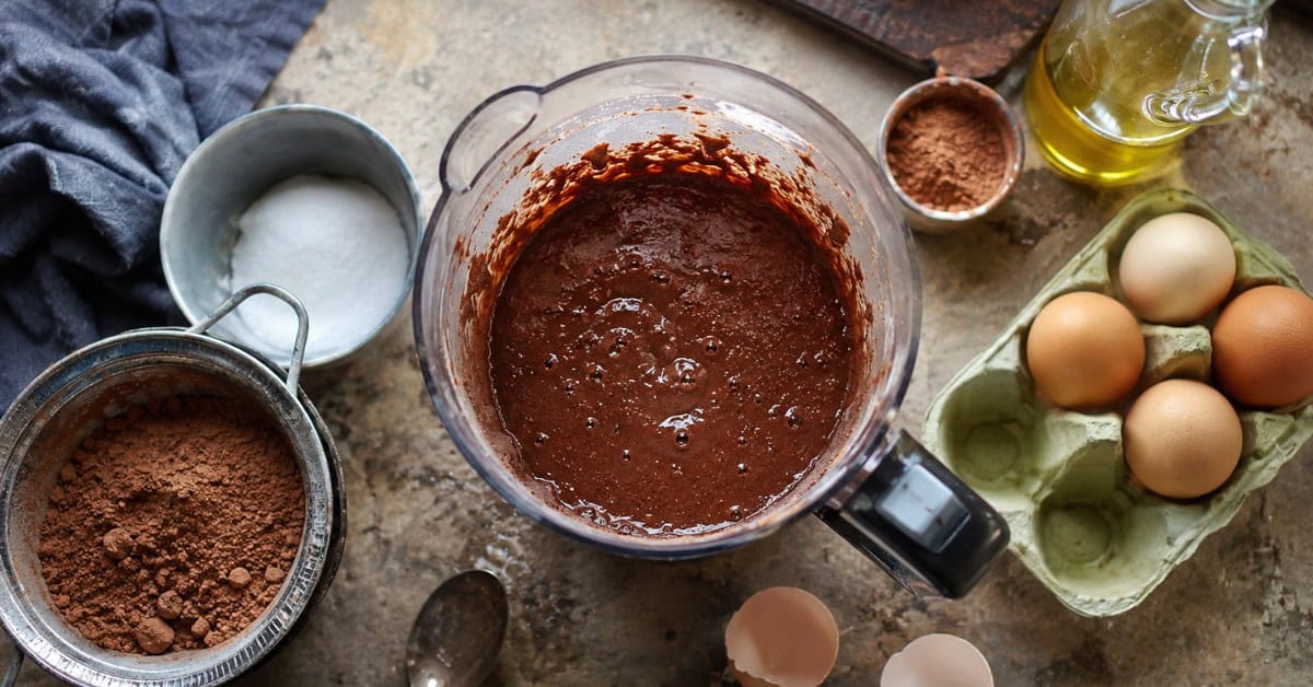 Liquidificador com massa de bolo de chocolate cercado por ingredientes como cacau em pó, ovos e adoçante sobre bancada de cozinha
