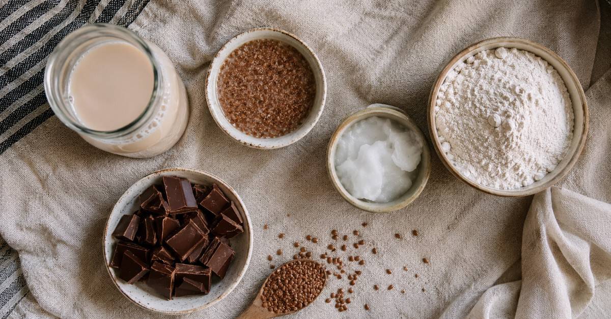 Pequenas tigelas com ingredientes substitutos para bolo sem açúcar: leite vegetal, farinha de arroz, óleo de coco, ovo de linhaça e chocolate zero açúcar sobre pano de linho