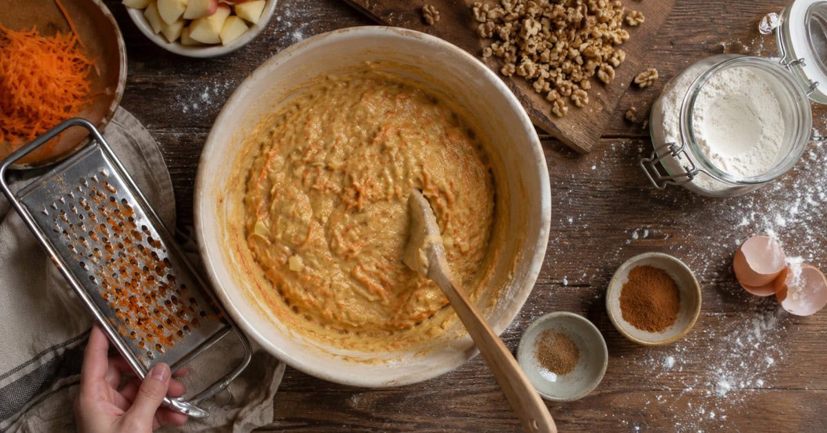 Tigela com massa laranja de cenoura e maçã raladas para bolo de nozes, com cenoura e utensílios ao redor