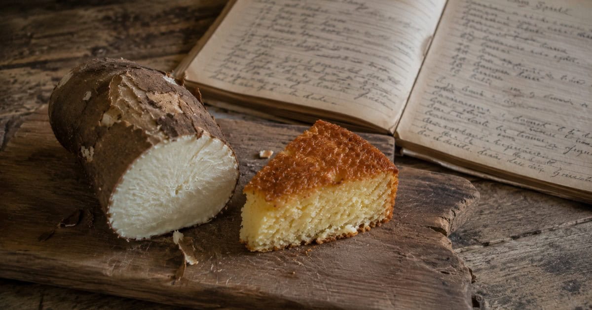 Tábua de madeira rústica com mandioca crua com casca ao lado de fatia de bolo assado e caderno de receitas manuscrito ao fundo