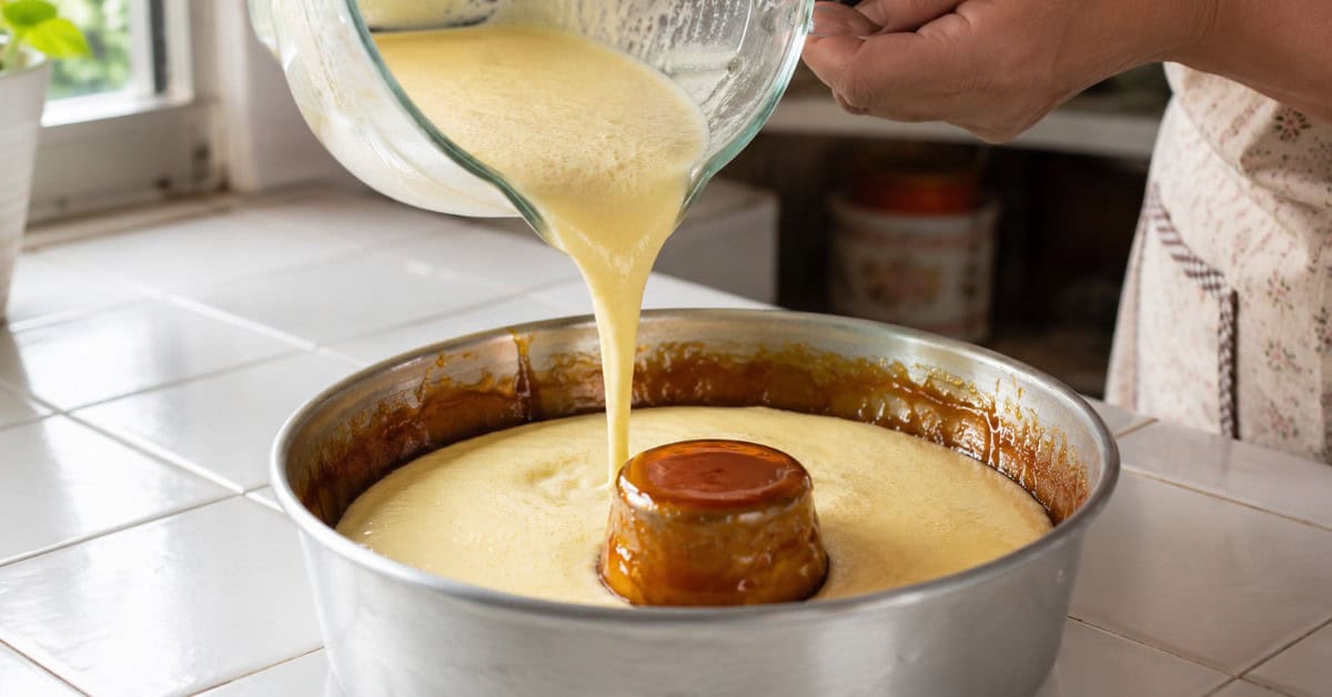 Mãos despejando creme de leite condensado e ovos na fôrma de pudim já caramelizada, em cozinha doméstica