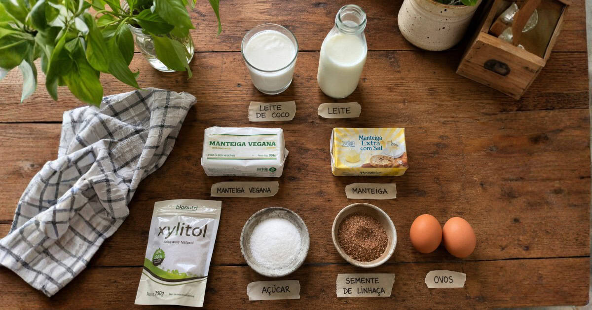 Flat lay com ingredientes substitutos para bolo de maisena sobre bancada rústica, incluindo leite de coco, manteiga vegetal, linhaça e xilitol lado a lado