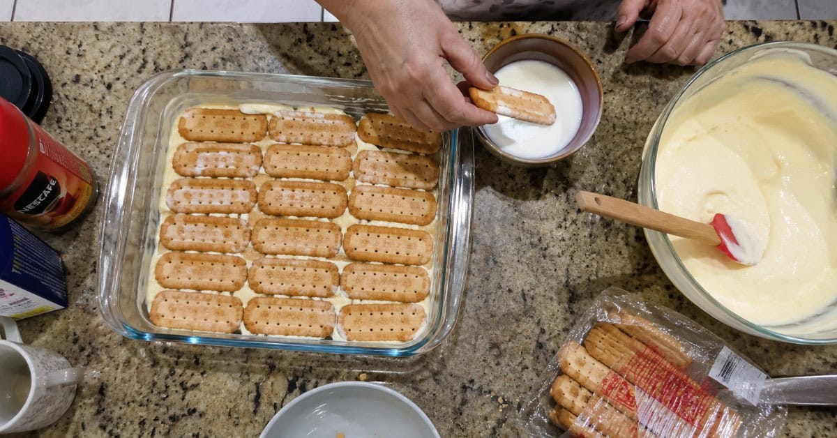 Mãos montando a torta alemã de leite ninho em refratário com camadas de biscoito e creme branco