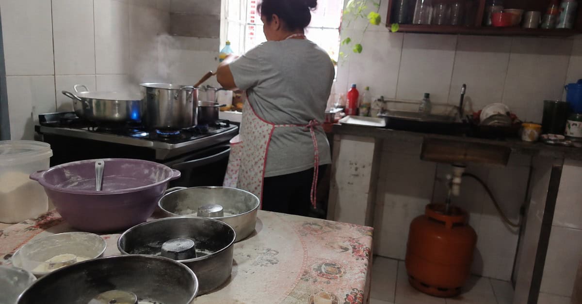 Mulher de avental trabalhando em cozinha doméstica com panelas no fogão aceso, forminhas de bolo na mesa enfarinhada e botijão de gás visível ao fundo