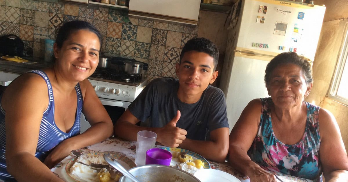 Família simples reunida na cozinha de casa com mãe, filho adolescente e avó à mesa com refeição e fogão a gás ao fundo