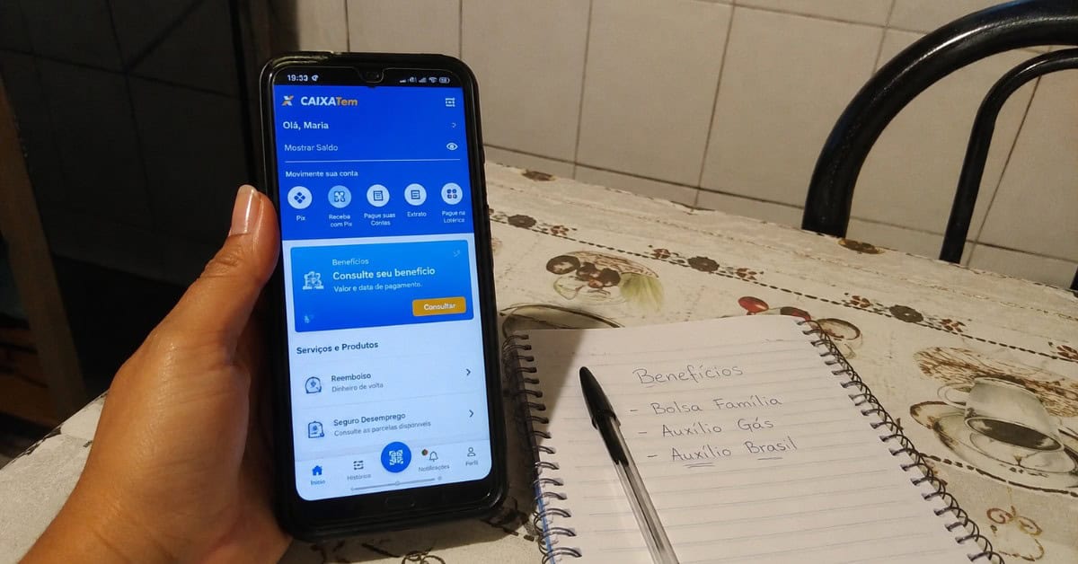 Mãos de mulher segurando celular com aplicativo Caixa Tem aberto sobre mesa de cozinha com caderno de anotações de benefícios sociais