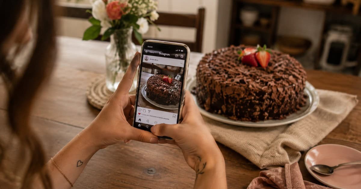 Mãos de mulher segurando celular fotografando bolo de chocolate caseiro sobre mesa com flores e tecido de linho, com Instagram aberto na tela