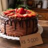 Bolo caseiro com ganache de chocolate e morangos frescos sobre mesa de madeira, com etiqueta escrita à mão mostrando R$ 5.000