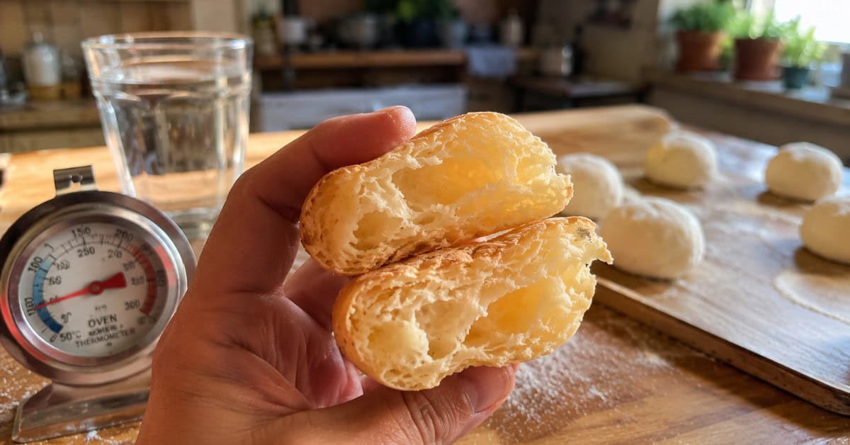 Mão segurando biscoito de polvilho partido ao meio mostrando o interior oco e macio com termômetro de forno ao fundo