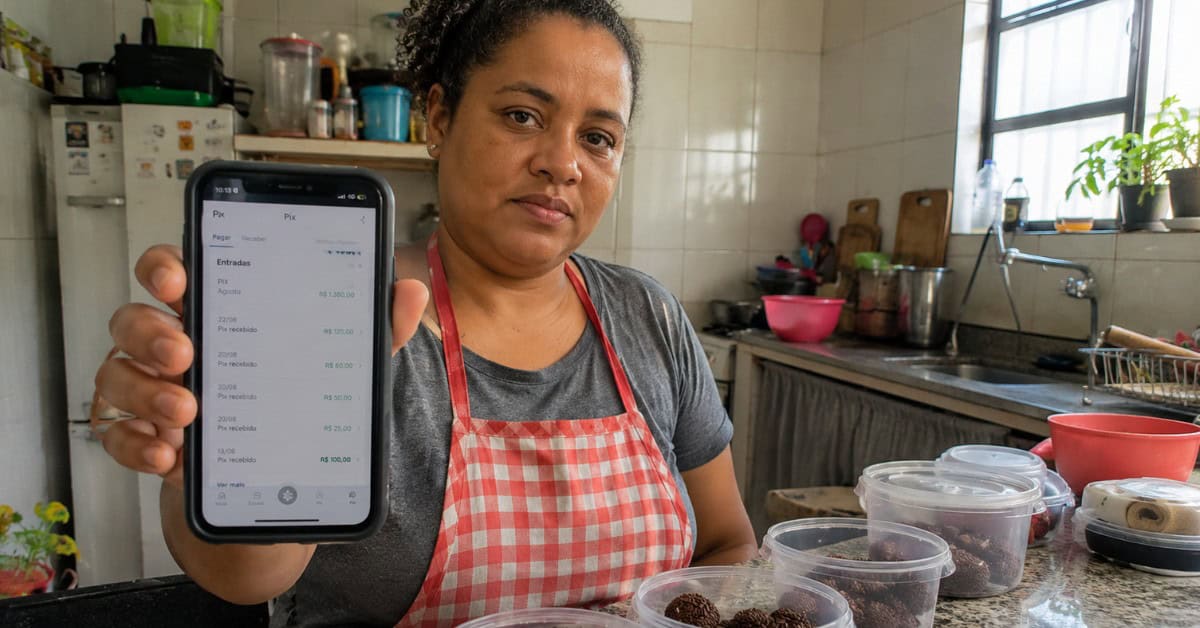 Mulher empreendedora em cozinha caseira mostrando extrato bancário no celular como comprovante de renda para o Minha Casa Minha Vida