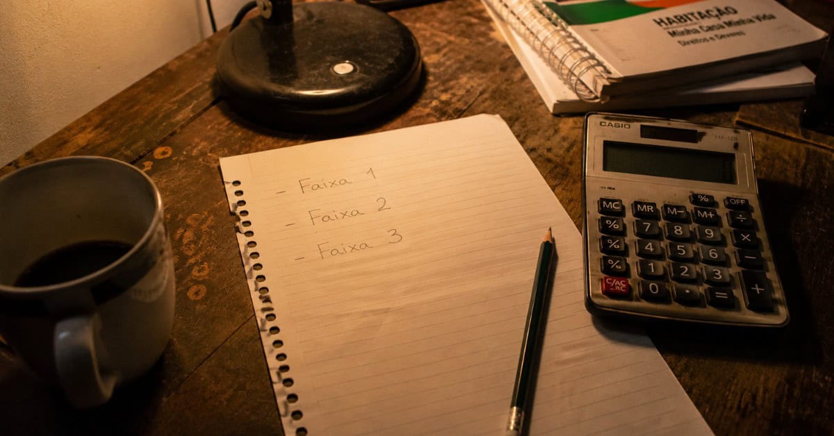 Papel com anotações à mão das faixas de renda do programa Minha Casa Minha Vida 2026 ao lado de uma calculadora e uma xícara de café