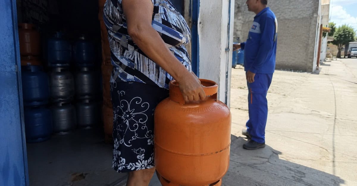 Mulher recebendo botijão de gás laranja de 13kg na porta de revenda credenciada com entregador ao fundo em bairro de periferia