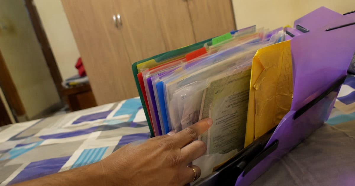 Pasta sanfonada com documentos para financiamento habitacional aberta sobre cama, com mão masculina apontando para os papéis do Minha Casa Minha Vida