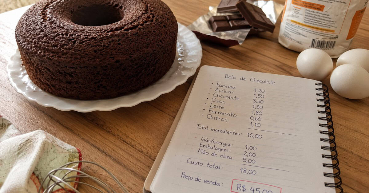 Bolo de chocolate caseiro sobre prato branco com caderno de custos aberto, ovos e barra de chocolate ao fundo, em cozinha iluminada por luz natural